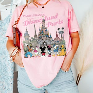 May include: Pink t-shirt featuring a Disneyland Paris graphic with the text "Paris, France Est. 1992". The graphic includes the Disneyland Paris castle and several Disney characters. The shirt is worn by a person.
