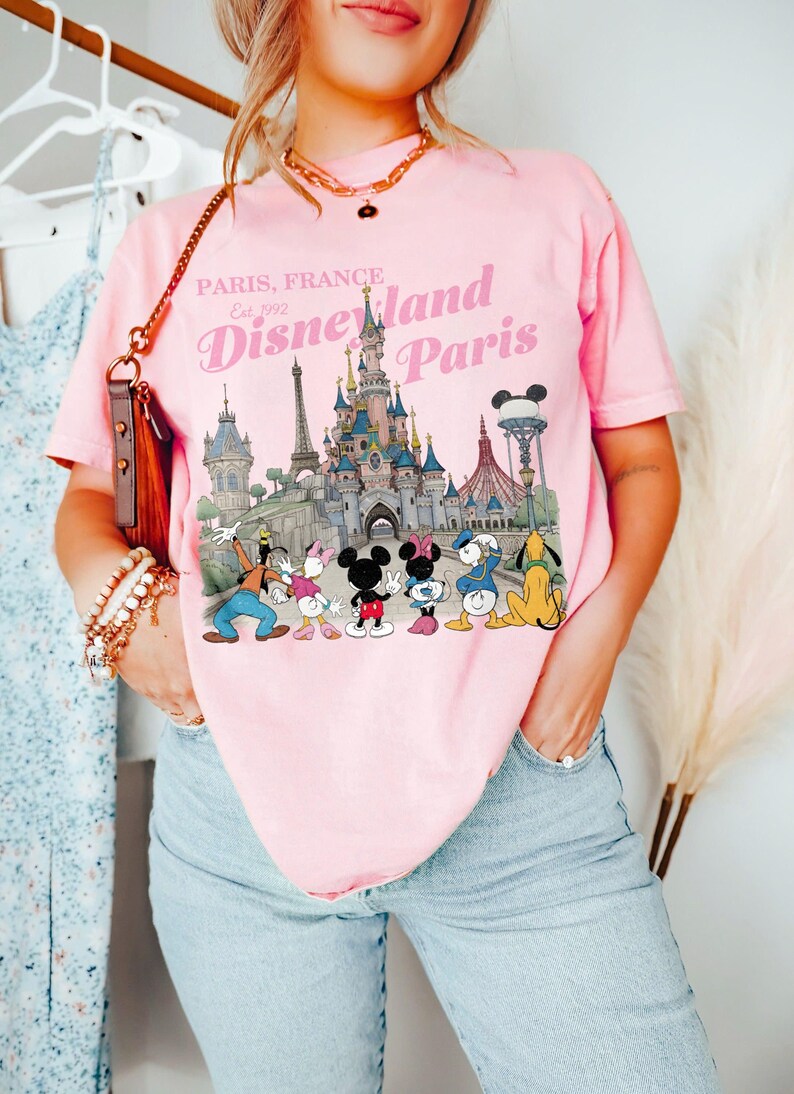 May include: Pink t-shirt featuring a Disneyland Paris graphic with the text "Paris, France Est. 1992". The graphic includes the Disneyland Paris castle and several Disney characters. The shirt is worn by a person.