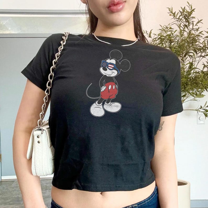 May include: A black short-sleeved t-shirt featuring a graphic print of a cartoon mouse wearing red shorts and sunglasses with a stars and stripes pattern. The design is centered on the chest of the shirt, which is paired with a white shoulder bag.