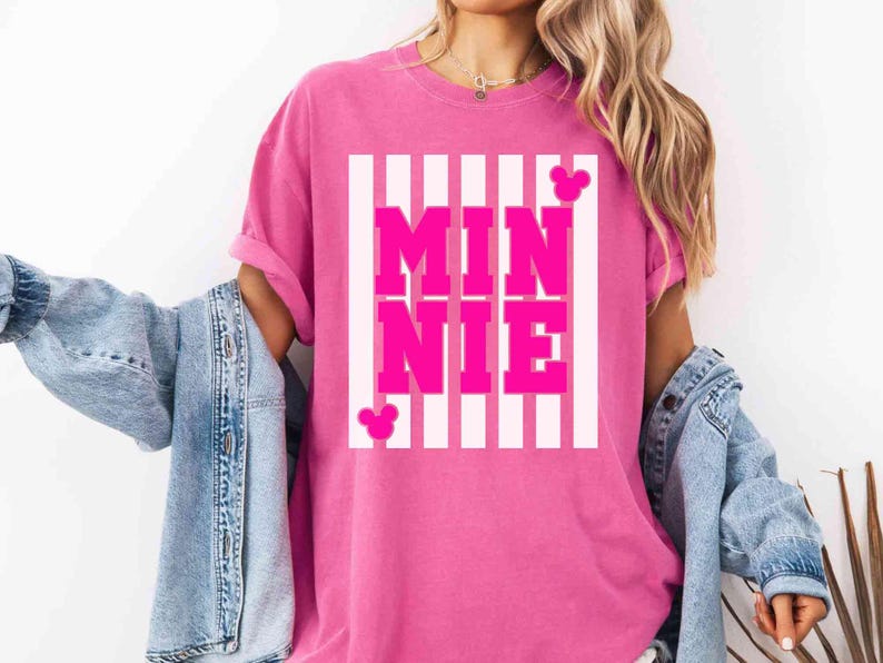 May include: A pink t-shirt with the name "MINNIE" in large, bold, pink letters, set against a white striped background. The shirt features two small Mickey Mouse head silhouettes. The shirt is a vibrant pink color.