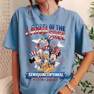 May include: Light blue t-shirt featuring a graphic of Disney characters in patriotic attire. The design includes the text "LAND OF THE FREE" and "SEMIQUINCENTENNIAL 1776-2026". The characters include Mickey and Minnie Mouse, Donald and Daisy Duck, Goofy, and Pluto.