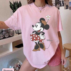 May include: A light pink t-shirt featuring a graphic of Minnie Mouse in a red polka dot dress, brown shoes, and a flower in her hat. The Disney logo is visible. The shirt is paired with red and white checkered shorts.
