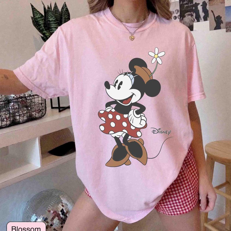 May include: A light pink t-shirt featuring a graphic of Minnie Mouse in a red polka dot dress, brown shoes, and a flower in her hat. The Disney logo is visible. The shirt is paired with red and white checkered shorts.
