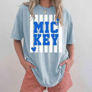 K&ouml;nnte beinhalten: Hellblaues T-Shirt mit dem Wort "MICKEY" in kr&auml;ftigen blauen Buchstaben, vor einem wei&szlig;-blauen Streifenhintergrund. Ein kleiner Mickey Mouse-Kopf befindet sich unter dem Text. Die Person tr&auml;gt rot-wei&szlig; karierte Shorts.