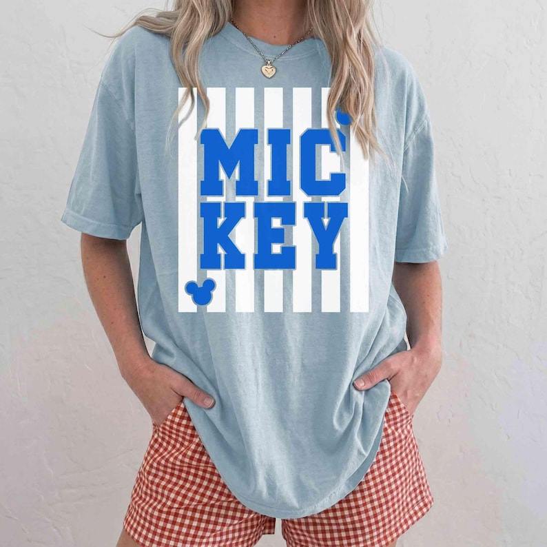 K&ouml;nnte beinhalten: Hellblaues T-Shirt mit dem Wort "MICKEY" in kr&auml;ftigen blauen Buchstaben, vor einem wei&szlig;-blauen Streifenhintergrund. Ein kleiner Mickey Mouse-Kopf befindet sich unter dem Text. Die Person tr&auml;gt rot-wei&szlig; karierte Shorts.