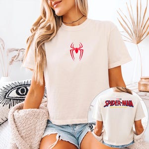 May include: Cream-colored t-shirt featuring a red spider symbol on the front. The back of the shirt, visible in a smaller inset image, displays the words "SPIDER-MAN" in red and blue.