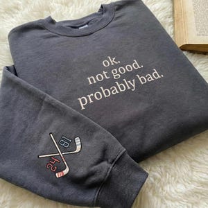 May include: Dark gray sweatshirt with the text "ok. not good. probably bad." in white. The left sleeve features crossed hockey sticks with the numbers 24 and 81. The sweatshirt is made of a soft material.