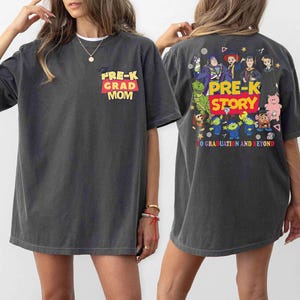 K&ouml;nnte beinhalten: Dunkelgraues T-Shirt mit "PRE-K GRAD MOM" in gelbem und rotem Text auf der Vorderseite. Die R&uuml;ckseite des Shirts zeigt eine farbenfrohe Grafik mit den Worten "PRE-K STORY" und verschiedenen Zeichentrickfiguren. Der Satz "TO GRADUATION AND BEYOND" ist ebenfalls auf der R&uuml;ckseite aufgedruckt.