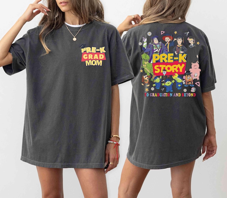 K&ouml;nnte beinhalten: Dunkelgraues T-Shirt mit "PRE-K GRAD MOM" in gelbem und rotem Text auf der Vorderseite. Die R&uuml;ckseite des Shirts zeigt eine farbenfrohe Grafik mit den Worten "PRE-K STORY" und verschiedenen Zeichentrickfiguren. Der Satz "TO GRADUATION AND BEYOND" ist ebenfalls auf der R&uuml;ckseite aufgedruckt.