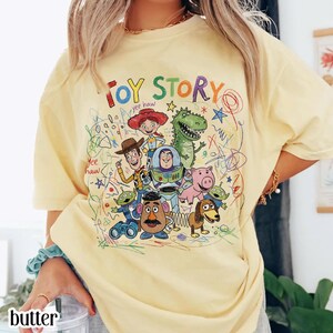 May include: A light yellow t-shirt featuring the words "TOY STORY" in colorful letters. The shirt showcases a vibrant graphic of characters from the animated film, including Woody, Buzz Lightyear, and Rex, with scribbled accents.
