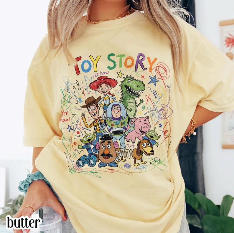 May include: A light yellow t-shirt featuring the words "TOY STORY" in colorful letters. The shirt showcases a vibrant graphic of characters from the animated film, including Woody, Buzz Lightyear, and Rex, with scribbled accents.