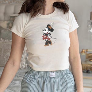 May include: A cream-colored short-sleeve t-shirt featuring a graphic of Minnie Mouse wearing a brown hat and a red polka-dot skirt, holding a white daisy. The word Disney is printed in a cursive script next to the character on the front.