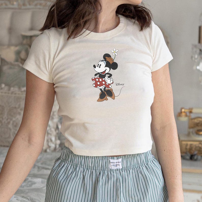 May include: A cream-colored short-sleeve t-shirt featuring a graphic of Minnie Mouse wearing a brown hat and a red polka-dot skirt, holding a white daisy. The word Disney is printed in a cursive script next to the character on the front.