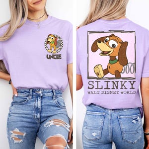 Puede incluir: Camiseta lila con una imagen de dibujos animados de Slinky Dog de Toy Story. La parte delantera muestra un gr&aacute;fico circular con la palabra "UNCLE". La parte trasera tiene un gr&aacute;fico rectangular con "SLINKY" y "WALT DISNEY WORLD".