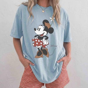 May include: Light blue t-shirt featuring a Minnie Mouse graphic. The cartoon character is wearing a red polka dot dress and brown shoes. The word "Disney" is printed in black script. The person is wearing red and white checkered shorts.