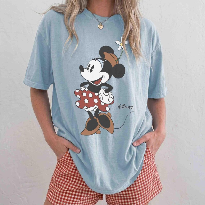 May include: Light blue t-shirt featuring a Minnie Mouse graphic. The cartoon character is wearing a red polka dot dress and brown shoes. The word "Disney" is printed in black script. The person is wearing red and white checkered shorts.