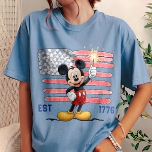 May include: Light blue t-shirt featuring Mickey Mouse holding a sparkler in front of a distressed American flag. The shirt has the text "EST 1776" in blue. The design is a patriotic theme.