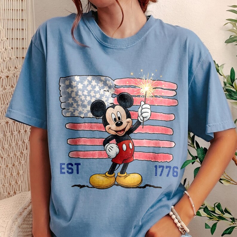 May include: Light blue t-shirt featuring Mickey Mouse holding a sparkler in front of a distressed American flag. The shirt has the text "EST 1776" in blue. The design is a patriotic theme.
