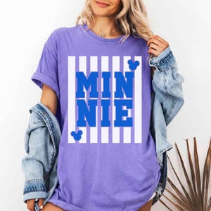 May include: A lilac-colored t-shirt with the word "MINNIE" in bold blue letters, set against a white striped background. Mickey Mouse silhouettes are placed above and below the text. The shirt has rolled-up sleeves.