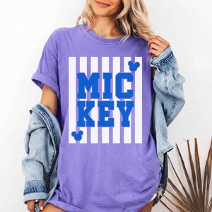 K&ouml;nnte beinhalten: Ein lila T-Shirt mit dem Wort "MICKEY" in blauen Blockbuchstaben, umrahmt von wei&szlig;en vertikalen Streifen. Mickey Mouse Silhouetten sind ebenfalls vorhanden. Das T-Shirt hat kurze &Auml;rmel und eine lockere Passform.