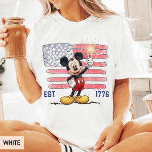 May include: White t-shirt featuring a graphic of Mickey Mouse holding a sparkler in front of an American flag design. The shirt has the text "EST 1776" in blue. The shirt is a casual style.