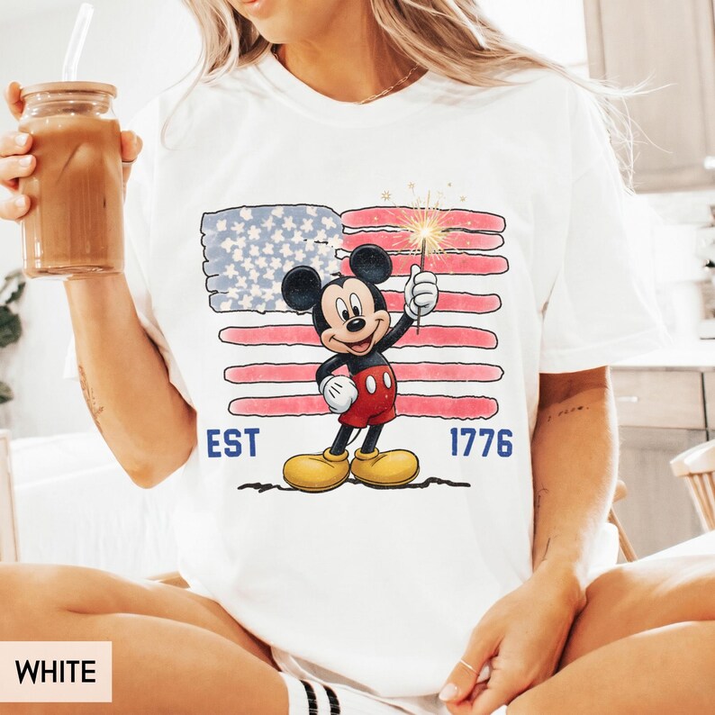 May include: White t-shirt featuring a graphic of Mickey Mouse holding a sparkler in front of an American flag design. The shirt has the text "EST 1776" in blue. The shirt is a casual style.