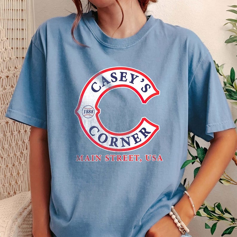May include: Light blue t-shirt with a distressed red and white graphic that reads "Casey's Corner" in a curved design. The text "Main Street, USA" is below the graphic. The shirt has short sleeves and a crew neck.