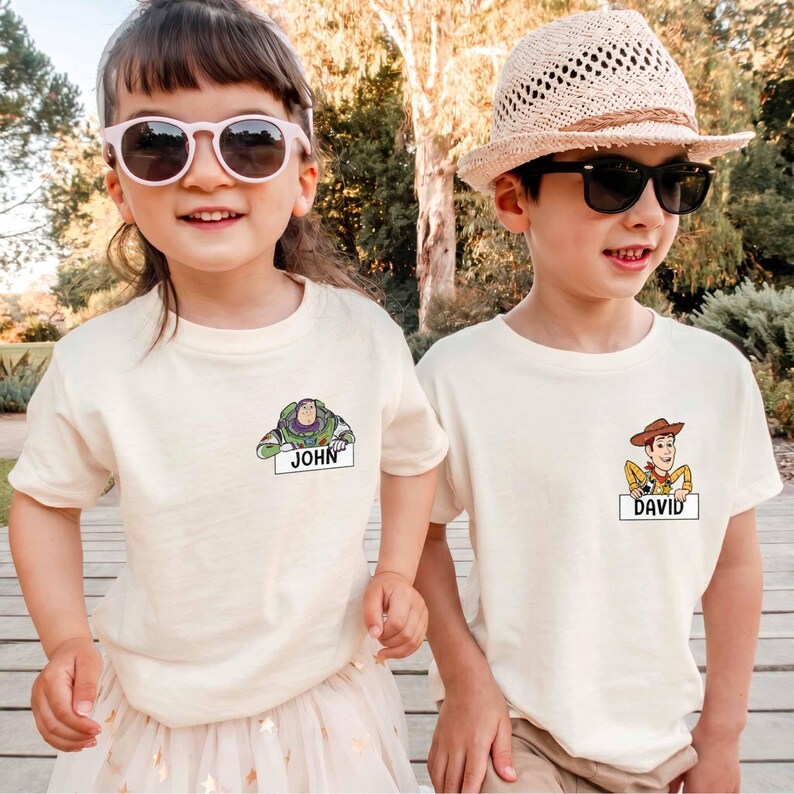 Peut inclure: Deux enfants portant des t-shirts blanc cass&eacute; avec des graphiques de personnages de dessins anim&eacute;s. Un t-shirt repr&eacute;sente Buzz l'&Eacute;clair avec le nom "JOHN", l'autre Woody avec le nom "DAVID". Les deux enfants portent des lunettes de soleil.