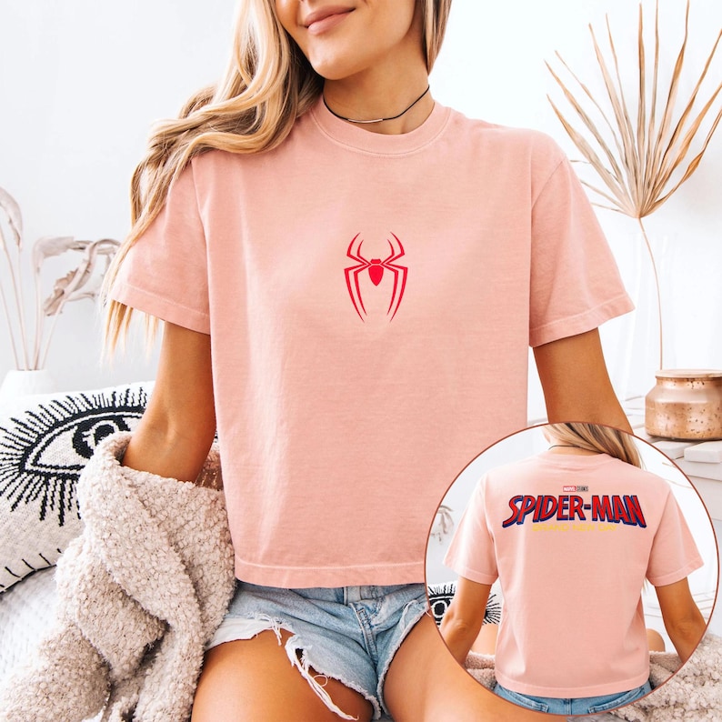 May include: A light pink t-shirt featuring a red spider emblem on the front. The back of the shirt displays the text "SPIDER-MAN" in a multi-colored design. The shirt is short-sleeved and has a crew neck.
