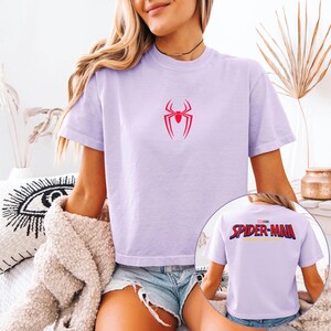 May include: A light purple t-shirt featuring a red spider logo on the front. The back of the shirt displays the text "SPIDER-MAN" in red, blue, and yellow. The shirt is short-sleeved and made of a soft material.