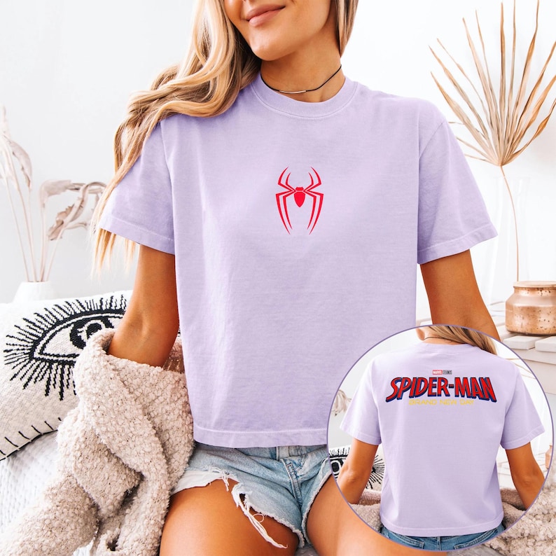 May include: A light purple t-shirt featuring a red spider logo on the front. The back of the shirt displays the text "SPIDER-MAN" in red, blue, and yellow. The shirt is short-sleeved and made of a soft material.