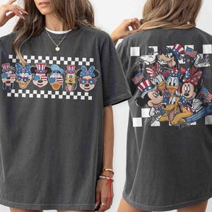 May include: Dark gray t-shirt featuring a patriotic design with cartoon characters. The front displays six character faces with American flag-themed accessories above a checkered pattern. The back showcases a group of characters holding flags. A fun, festive tee.