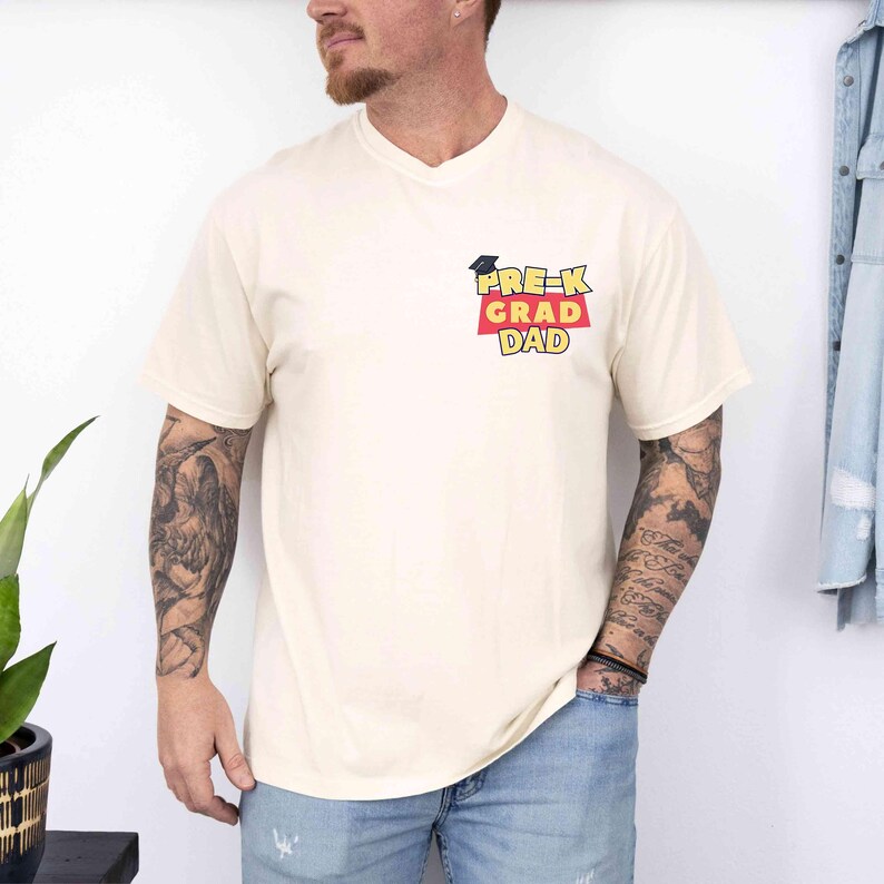 K&ouml;nnte beinhalten: Cremefarbenes T-Shirt mit V-Ausschnitt. Das Shirt hat einen kleinen Aufdruck auf der Brust mit der Aufschrift "PRE-K GRAD DAD" in Schwarz, Rot und Gelb. Eine Abschlusskappe befindet sich &uuml;ber dem Text. Das Shirt ist aus weichem Material.