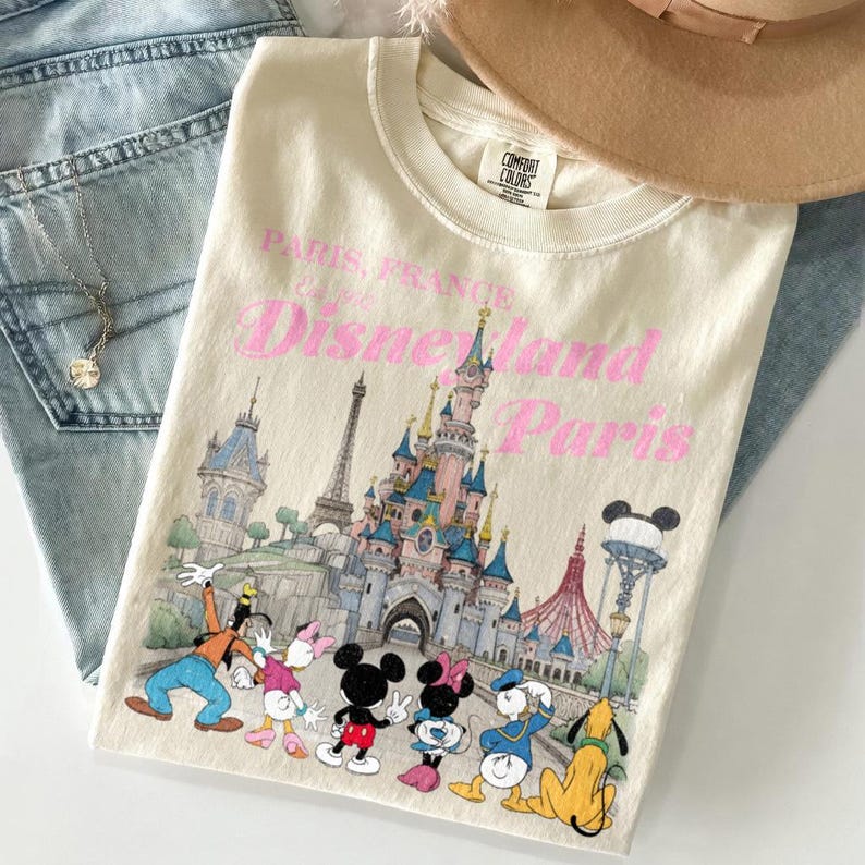 May include: Cream-colored t-shirt featuring a Disneyland Paris graphic. The design includes the Eiffel Tower, a castle, and various Disney characters. The text "Paris, France Disneyland Paris" is printed in pink.