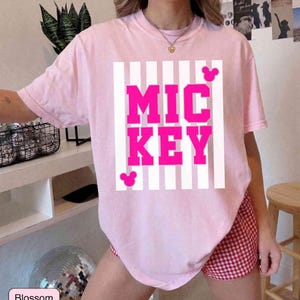 K&ouml;nnte beinhalten: Ein hellrosa T-Shirt mit dem Wort "MICKEY" in leuchtend pinkfarbenen Blockbuchstaben, vor einem wei&szlig; gestreiften Hintergrund. Das Shirt hat zwei kleine Mickey Mouse Kopf-Silhouetten. Das Wort "Blossom" steht in einer kleinen Box unten.