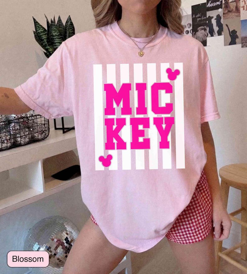 K&ouml;nnte beinhalten: Ein hellrosa T-Shirt mit dem Wort "MICKEY" in leuchtend pinkfarbenen Blockbuchstaben, vor einem wei&szlig; gestreiften Hintergrund. Das Shirt hat zwei kleine Mickey Mouse Kopf-Silhouetten. Das Wort "Blossom" steht in einer kleinen Box unten.