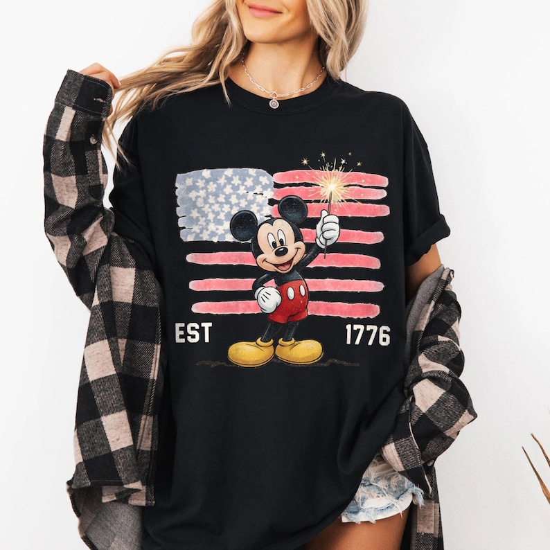 May include: Black t-shirt featuring Mickey Mouse holding a sparkler in front of a stylized American flag. The design includes the text "EST 1776". Mickey Mouse is wearing red shorts, yellow shoes, and white gloves.