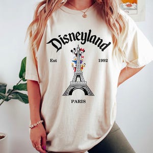 May include: Cream-colored t-shirt featuring a black graphic of the Eiffel Tower with Disney characters. The word "Disneyland" is arched above the tower, with "Est 1992" on either side. The word "Paris" is below the tower.