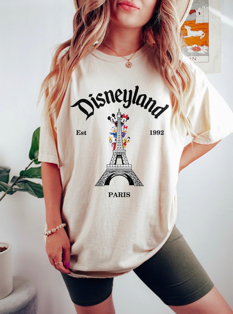 May include: Cream-colored t-shirt featuring a black graphic of the Eiffel Tower with Disney characters. The word "Disneyland" is arched above the tower, with "Est 1992" on either side. The word "Paris" is below the tower.