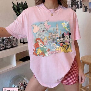 May include: A light pink t-shirt featuring a colorful graphic of Disney characters, including Dumbo, Ariel, and Mickey Mouse, in front of a castle. The word "Disneyland" is printed below the characters. The shirt is paired with red and white checkered shorts.