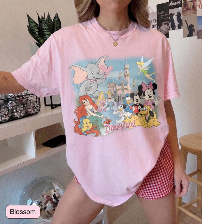 May include: A light pink t-shirt featuring a colorful graphic of Disney characters, including Dumbo, Ariel, and Mickey Mouse, in front of a castle. The word "Disneyland" is printed below the characters. The shirt is paired with red and white checkered shorts.