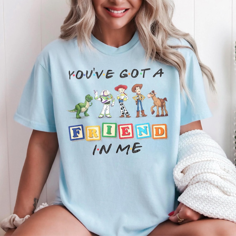May include: A light blue t-shirt featuring a graphic of five animated characters standing in a row above colorful alphabet blocks that spell out the word FRIEND. The text YOU'VE GOT A is printed above the characters and IN ME is printed below.