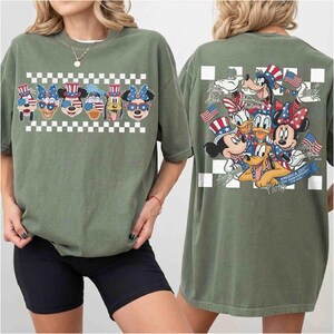 May include: Olive green t-shirt featuring a patriotic design with Disney characters. The front displays character faces with American flag-themed accessories above a checkered pattern. The back showcases a group of characters with flags and text.