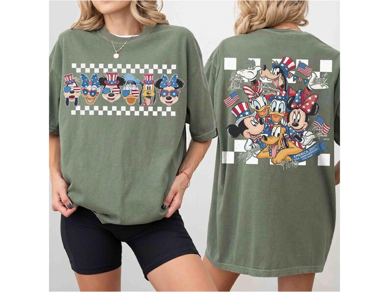 May include: Olive green t-shirt featuring a patriotic design with Disney characters. The front displays character faces with American flag-themed accessories above a checkered pattern. The back showcases a group of characters with flags and text.