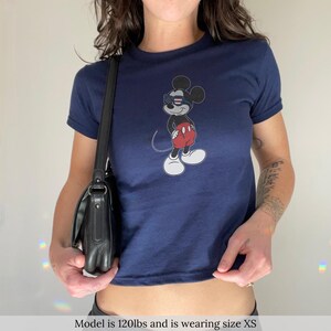 May include: A navy blue short-sleeved t-shirt featuring a graphic of a cartoon mouse wearing red shorts and sunglasses with a red, white, and blue star-spangled pattern. The shirt is paired with a black leather shoulder bag with a silver buckle.
