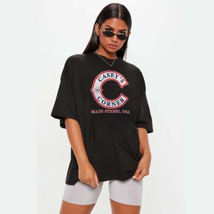 May include: Black oversized t-shirt with a large graphic of "Casey's Corner" in red, white, and blue lettering. The shirt features a classic collegiate-style design and the words "Main Street, USA". The model is wearing gray shorts.