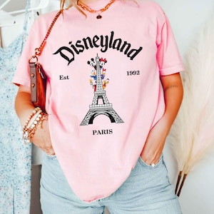 May include: Pink t-shirt with a Disneyland Paris graphic. The design features the Eiffel Tower with Disney characters and the text "Disneyland Est 1992 Paris". The shirt is a light pink color.