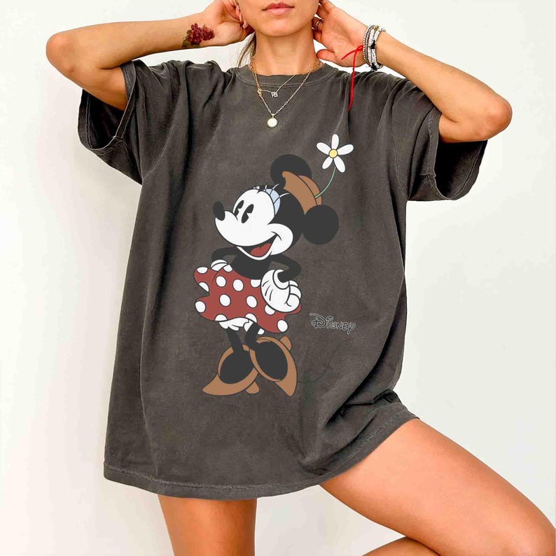 May include: Dark grey t-shirt featuring a Minnie Mouse graphic. Minnie is wearing a red polka dot dress, brown shoes, and a hat with a white flower. The Disney logo is visible on the bottom right.