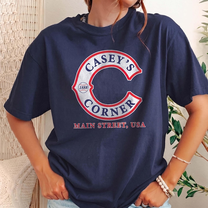 May include: Navy blue t-shirt featuring the text "Casey's Corner" in a white and red design. The design includes the text "Main Street, USA" below the logo. The shirt has short sleeves and a crew neck.