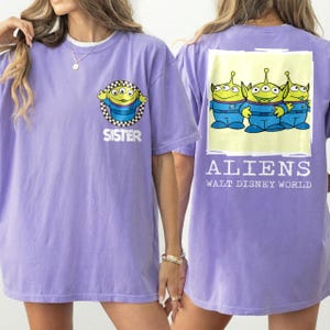 Puede incluir: Camiseta de color lila con un alien&iacute;gena de dibujos animados en la parte delantera y la palabra "SISTER". La parte trasera muestra tres personajes alien&iacute;genas y el texto "ALIENS WALT DISNEY WORLD".